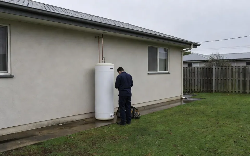 A plumber in a navy work shirt at an electric storage hot water cylinder against the rendered side wall of a house, on an overcast day
